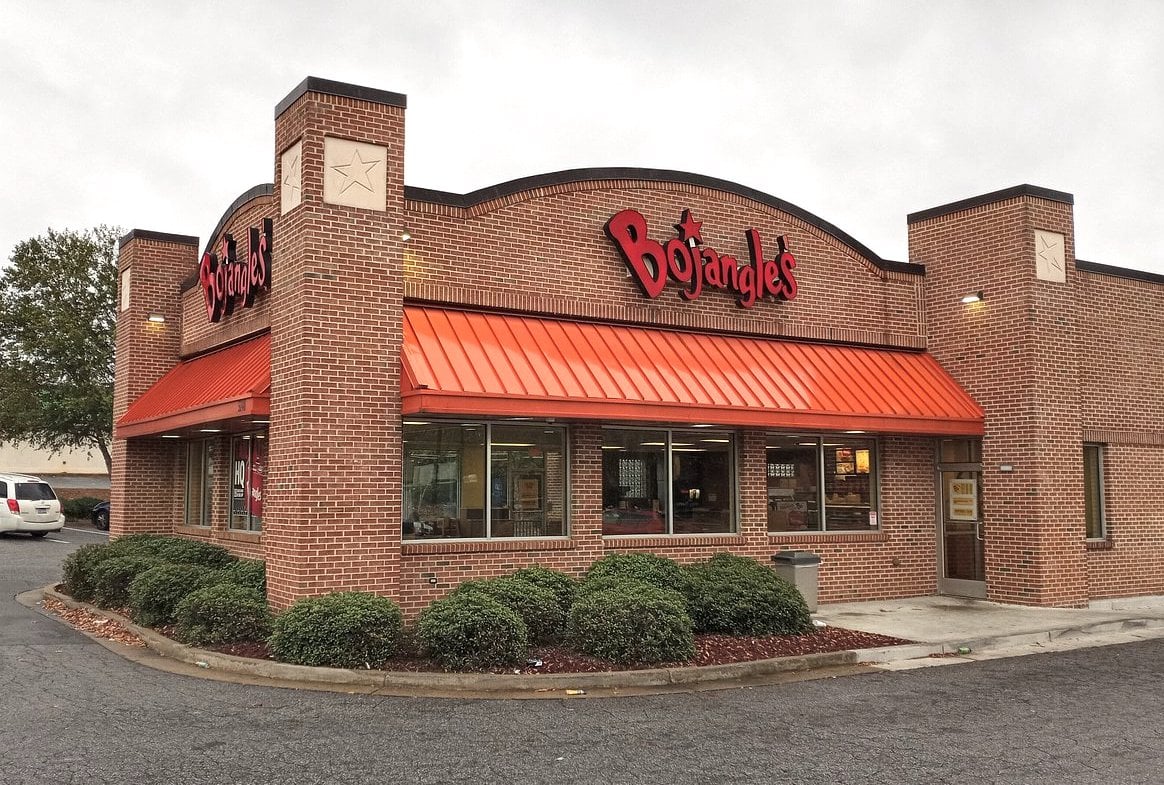 A Bojangles restaurant with a red-orange metal roof, red brick exterior, and a large red Bojangles sign above the entrance, surrounded by shrubs and a parking lot on a cloudy day.
