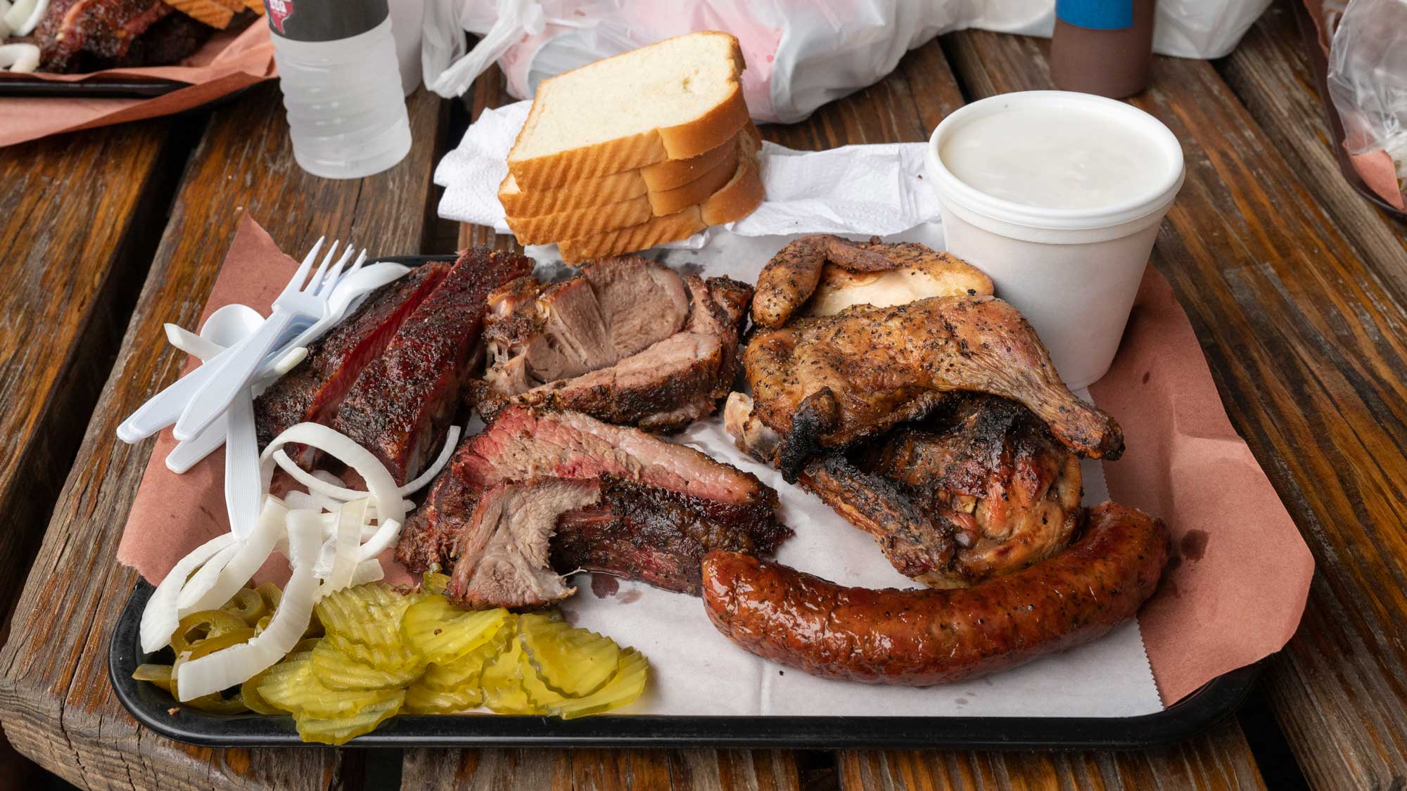 A tray of the best BBQ food with sliced brisket, sausage, smoked chicken, pickles, onions, sliced white bread, plastic utensils, and a cup of white sauce on a wooden table.