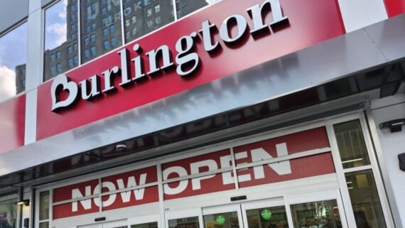 Storefront of Burlington with large red and white sign above the entrance, and a "NOW OPEN" banner displayed across the glass doors. Reflective windows show surrounding buildings.