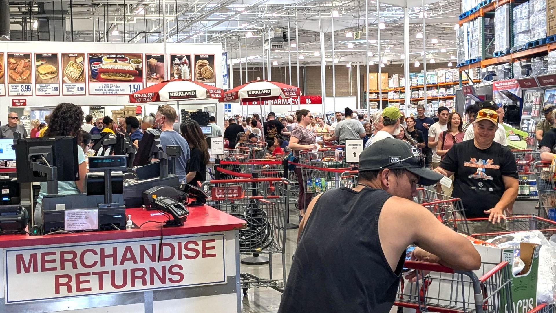 A busy wholesale Costco store with many people in line at the merchandise returns counter. Shopping carts are filled with goods, and signs and products are visible in the background. The atmosphere is crowded and bustling.