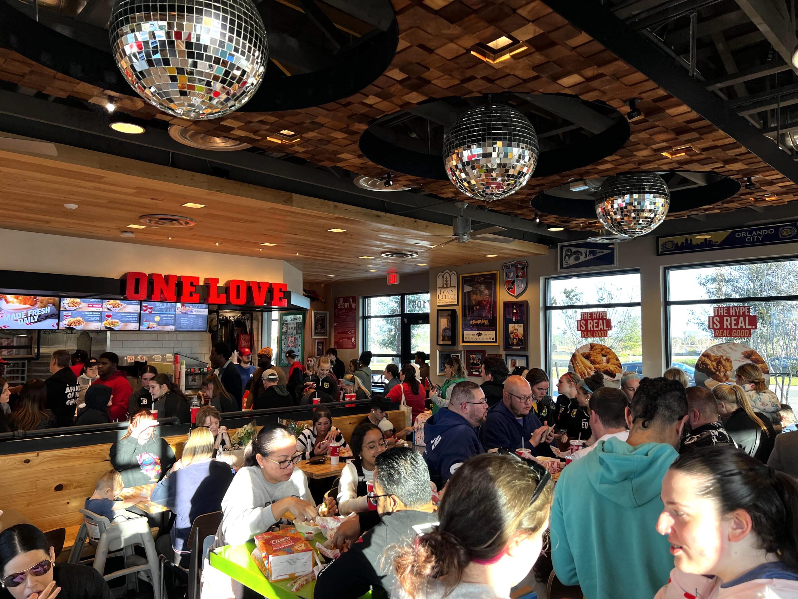 A crowded fast-food restaurant with many people sitting at tables and standing in line. Bright sunlight streams through the windows, and disco balls hang from the ceiling. A sign on the wall reads "ONE LOVE.