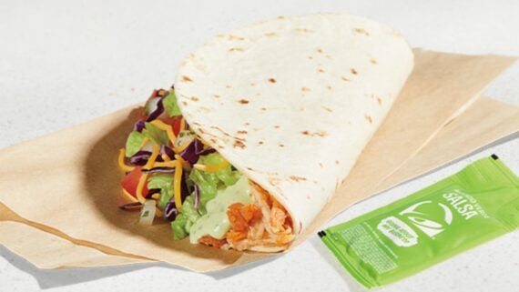A soft tortilla wrap filled with shredded chicken, lettuce, cheese, and tomatoes on brown paper, next to a green packet of mild salsa.
