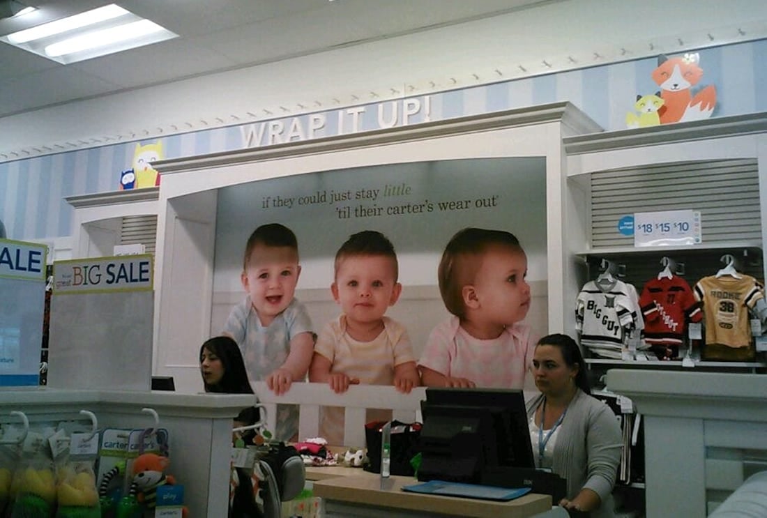 A retail store interior with two employees at the counter, a large poster of three babies above them, and baby clothes on display. The sign above reads "WRAP IT UP!" with a sale section visible on the left.