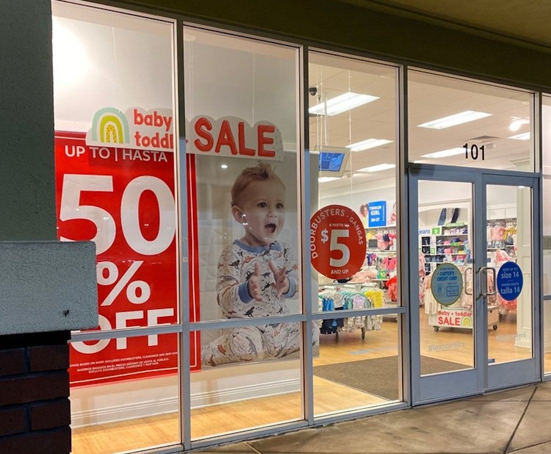 A brightly lit store window displays large signs advertising a sale with "50% OFF" and "Busters $5 and up," alongside a large image of a baby in pajamas. The store interior with baby items is visible inside.