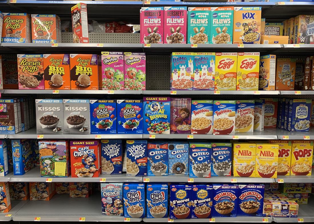 Grocery store shelves stocked with various colorful cereal boxes, including brands like Reese’s Puffs, Froot Loops, Eggo, Oreo O’s, Cap’n Crunch, and Pops, arranged in rows.