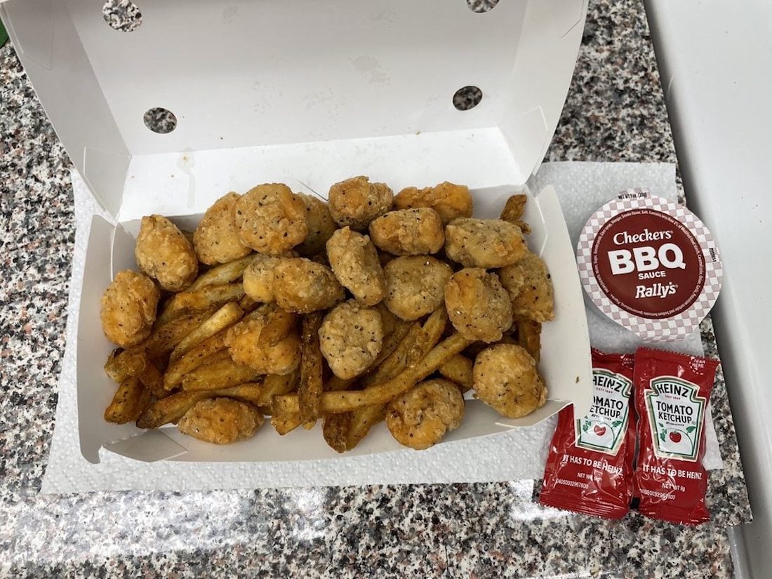 An open takeout box with seasoned fries and chicken nuggets sits on a counter. Next to it are a cup of Checkers Rally's BBQ sauce and two packets of Heinz tomato ketchup.