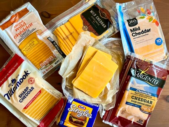 Various packages of sliced cheddar cheese, both mild and sharp, from different brands are arranged on a wooden surface, with some cheese slices unwrapped in the center.