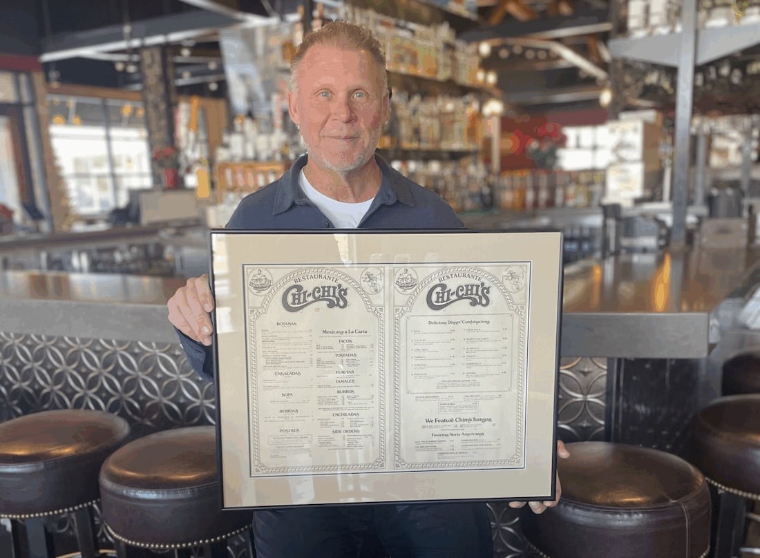 A man with short light hair and a blue shirt stands in a modern restaurant, smiling and holding a large framed menu with ornate lettering and two columns of vintage-style text. Barstools and shelves are visible in the background.