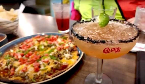 A plate of nachos topped with meat, cheese, and tomatoes sits next to a margarita garnished with a lime and a salt rim on a restaurant table.
