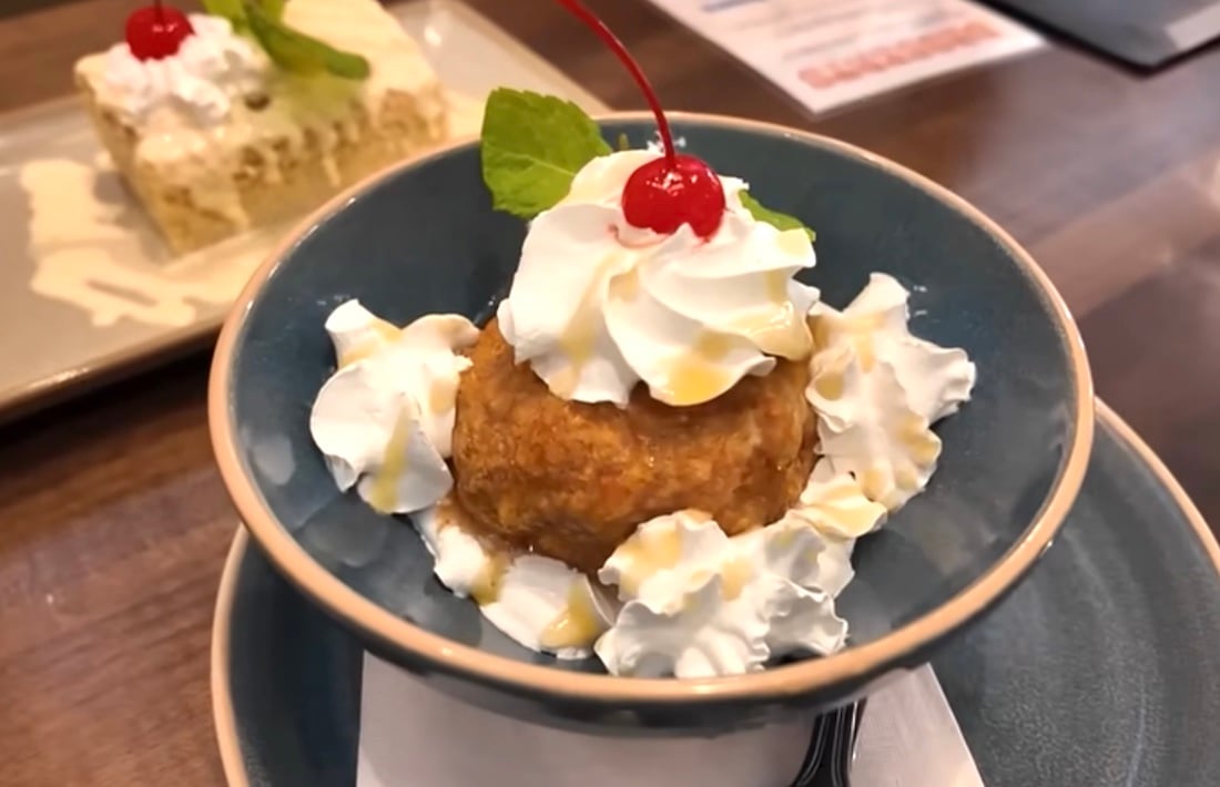 A bowl of fried ice cream topped with whipped cream, caramel drizzle, a maraschino cherry, and a mint leaf, with more whipped cream around it. A fork and a dessert plate are in the background.