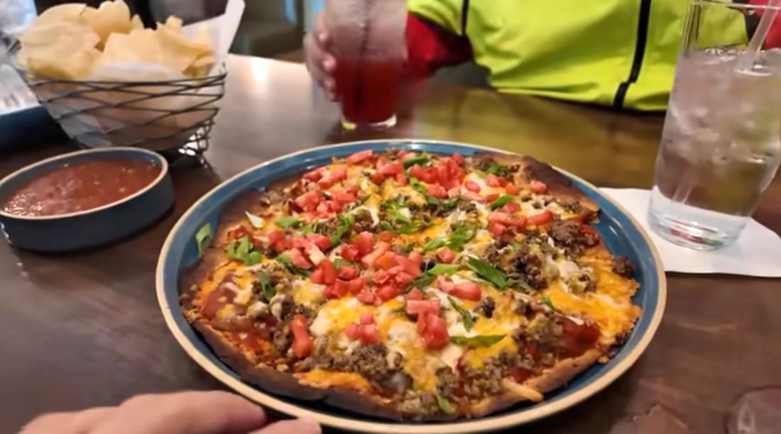 A plate of nachos topped with ground beef, melted cheese, chopped tomatoes, and green onions sits on a wooden table beside a bowl of salsa, a basket of tortilla chips, and two drinks—one clear and one red.