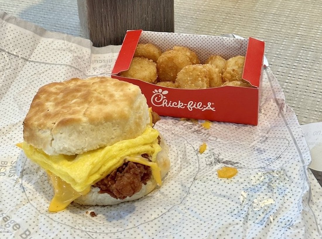 A Chick-fil-A biscuit sandwich with egg, cheese, and chicken sits on a wrapper next to a small red Chick-fil-A box filled with round hash browns.