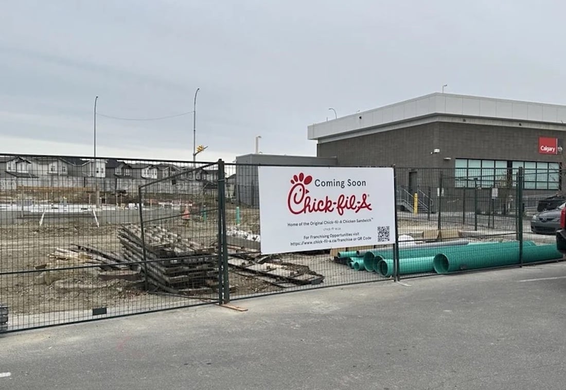 A fenced-off construction site with a sign reading "Coming Soon Chick-fil-A" in front of an unfinished building. Pipes and construction materials are visible on the ground.
