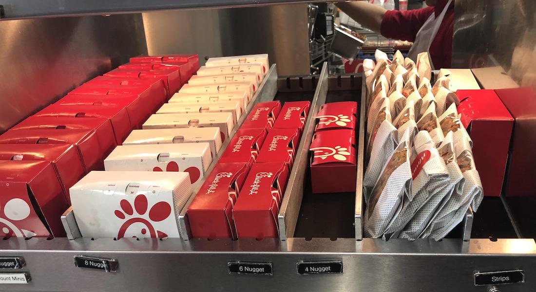 A row of Chick-fil-A boxes and bags filled with food items, such as nuggets and sandwiches, organized on a metal warming shelf at a restaurant counter.