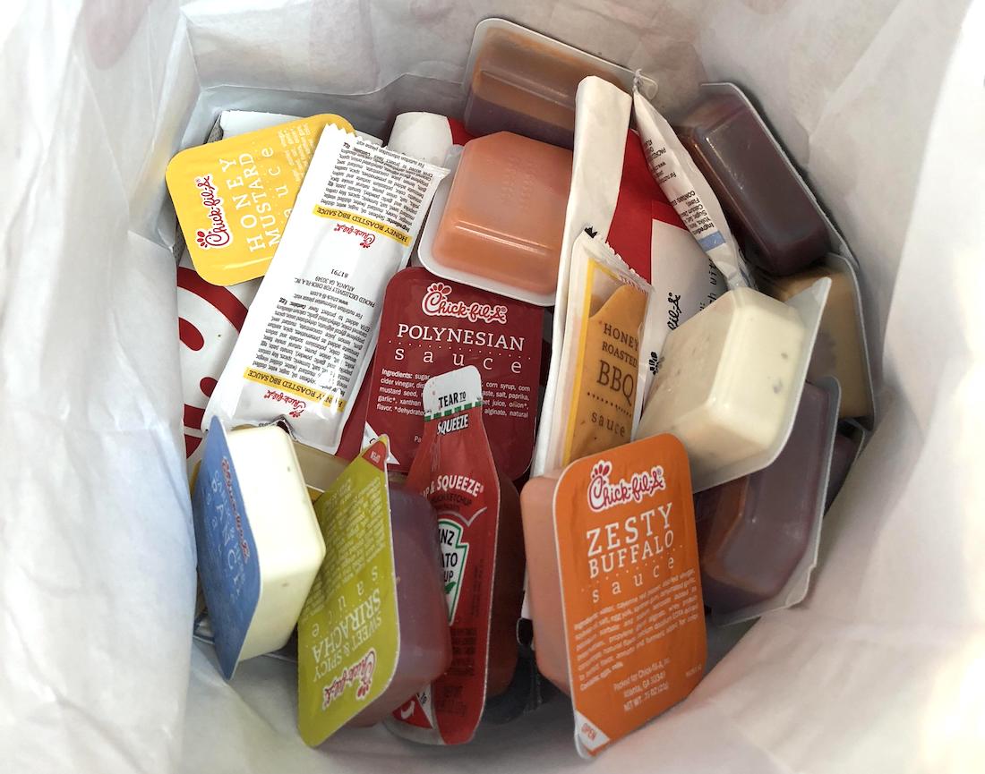 A collection of assorted fast food sauce packets, including Chick-fil-A sauces, ketchup, and ranch, inside a white paper bag.