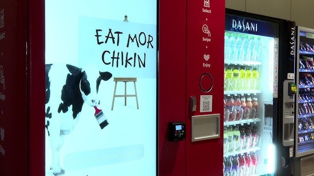 A vending machine with a display showing a cow holding a paintbrush, painting the words "EAT MOR CHIKIN" on a sign, next to another vending machine filled with various drinks.