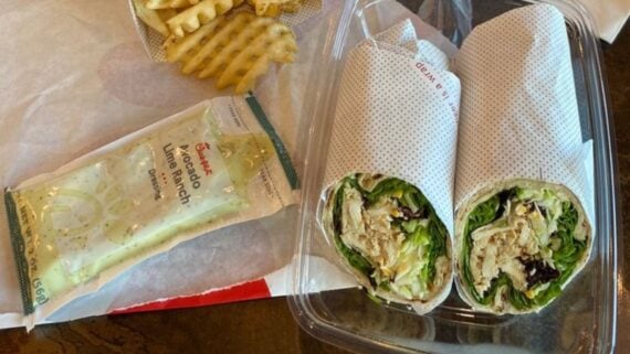 A plastic container holds a wrap sliced in half, filled with lettuce and chicken. Beside it are waffle fries on a tray and a packet of Chick-fil-A avocado lime ranch dressing.