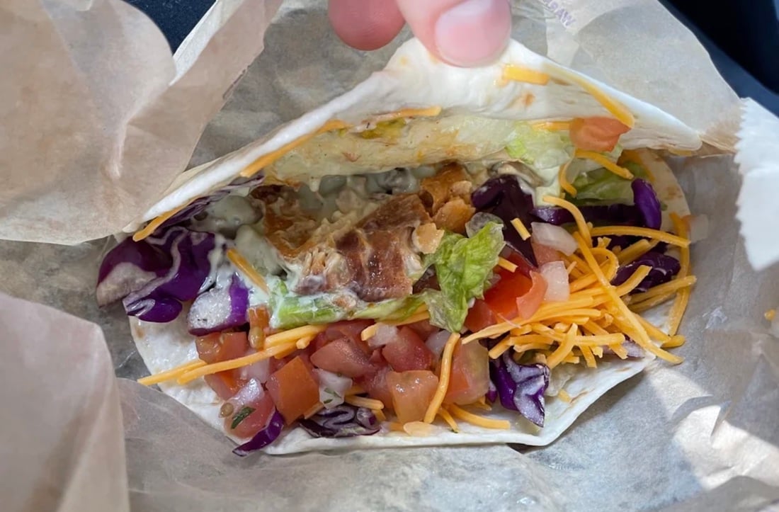 A close-up of a hand holding an open soft taco filled with shredded cheese, diced tomatoes, lettuce, purple cabbage, grilled chicken, and sauce in a flour tortilla.