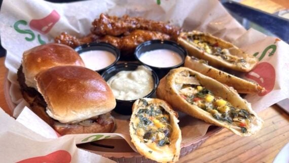 A platter with two sliders, crispy egg rolls stuffed with vegetables, saucy chicken tenders, and three cups of dipping sauce, all served on branded paper in a basket.