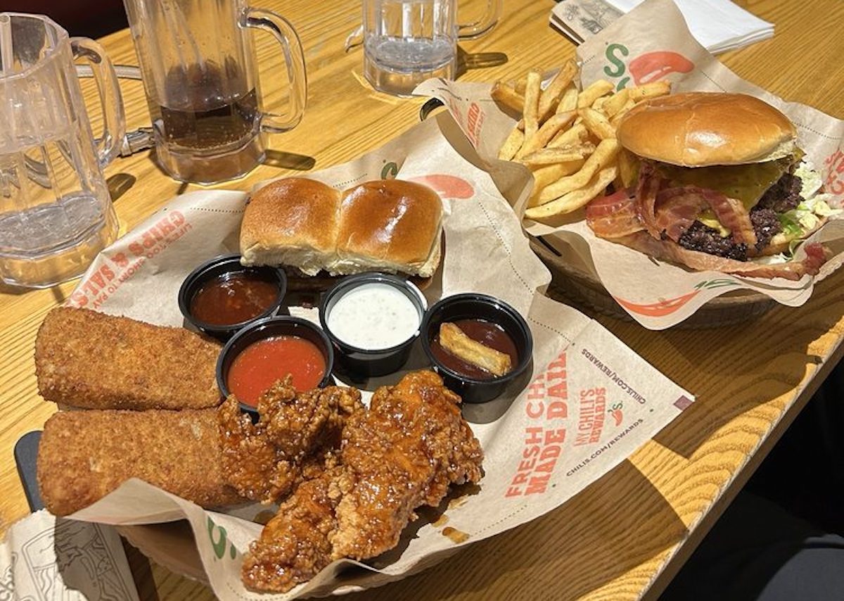 A restaurant table with mozzarella sticks, chicken tenders with sauces, two bread rolls, a burger with lettuce and bacon, fries, and empty glass mugs. Food is served on branded paper.