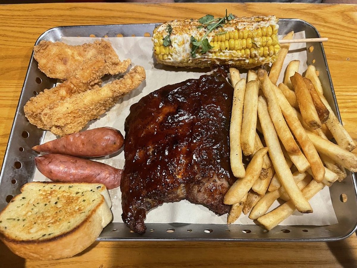 A tray with fried chicken tenders, two sausages, BBQ ribs, French fries, grilled corn on the cob with sauce and herbs, and a slice of garlic bread on parchment paper.