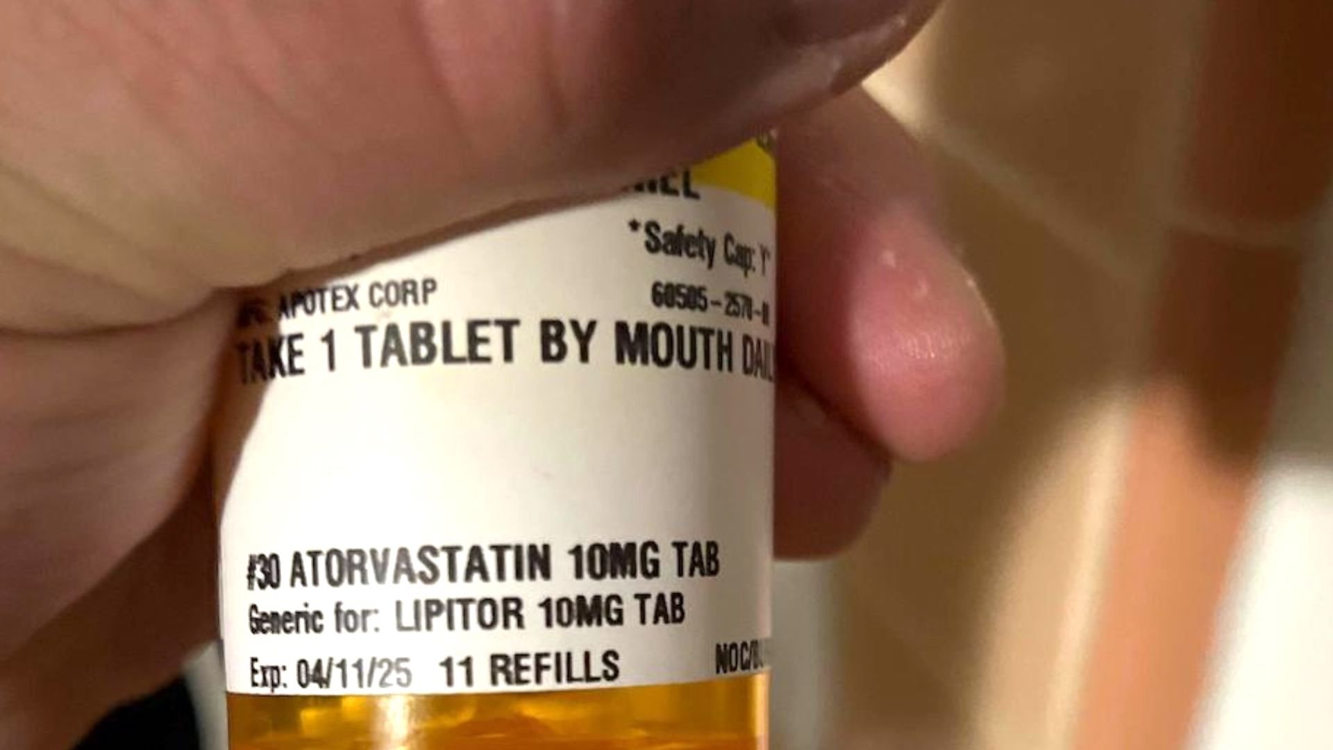 A close-up of a person's hand holding a prescription bottle labeled "Atorvastatin 10mg tab," with instructions to take one tablet by mouth daily. The label mentions "Generic for: Lipitor 10mg tab" and shows 11 refills.
