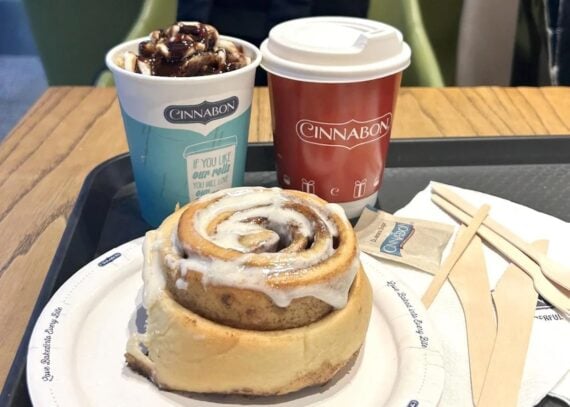 A Cinnabon cinnamon roll with icing is on a plate, next to two Cinnabon cups—one with a hot drink and the other with what looks like an iced dessert topped with chocolate sauce—on a tray with napkins and wooden stirrers.