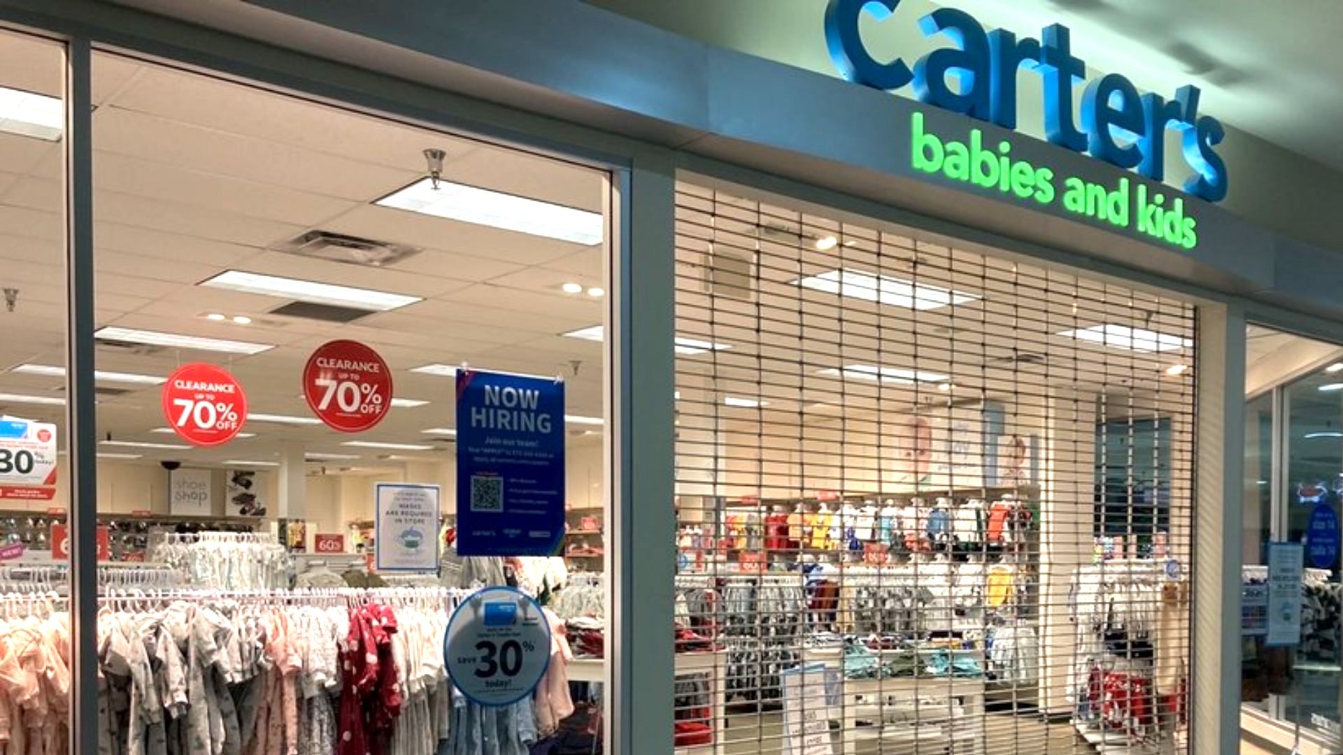 Storefront of Carter’s babies and kids, with a metal gate partially down. Sale signs advertising 70% off and a “Now Hiring” sign are visible. Racks of children’s clothing are displayed inside the store.