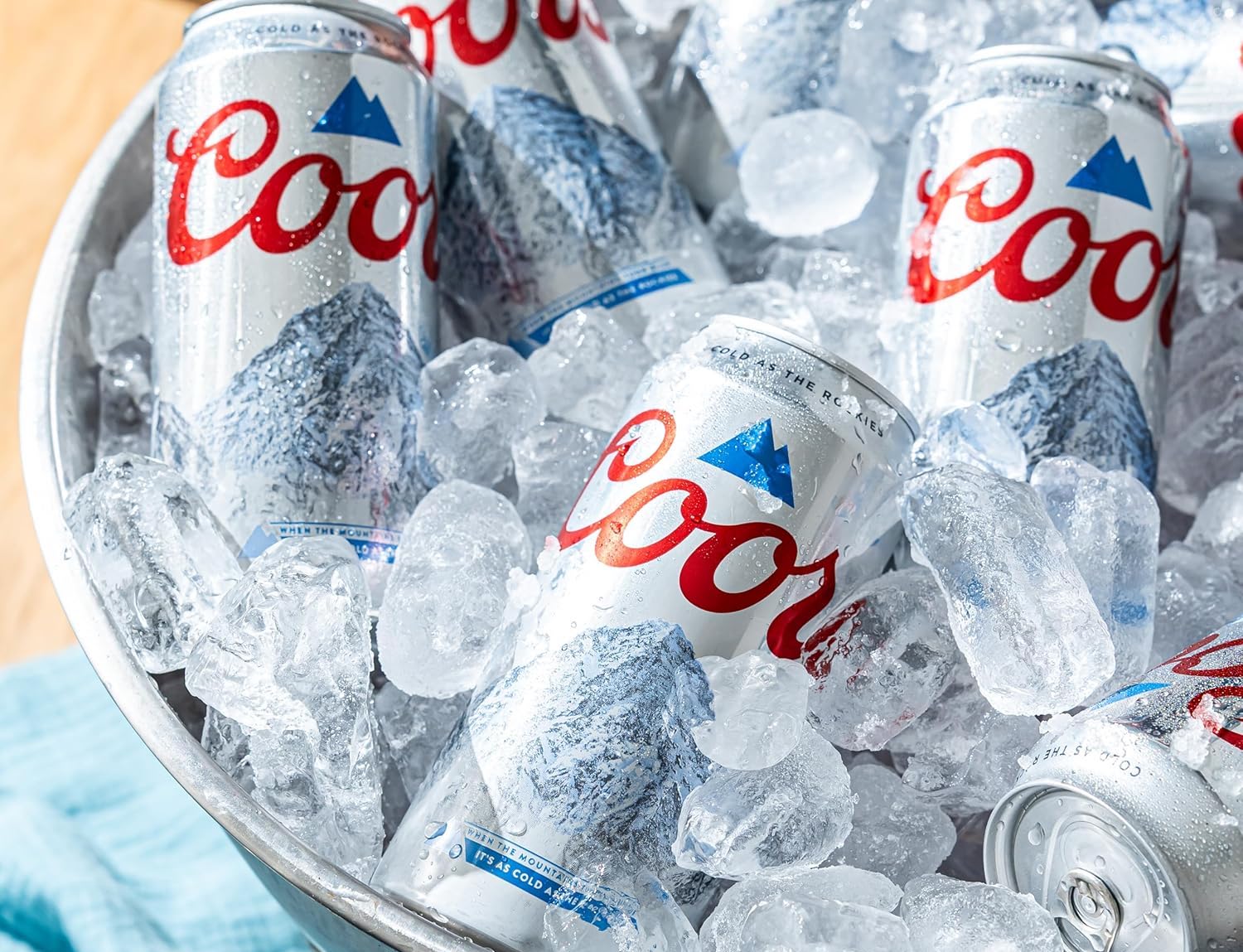 Several cans of Coors beer are chilled in a large metal bucket filled with ice cubes, with condensation visible on the cans and ice.