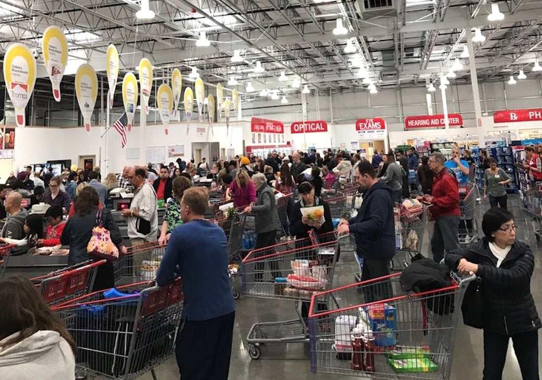 A crowded warehouse store with many people waiting in long checkout lines, pushing shopping carts filled with groceries and household items under bright lights and high ceilings.