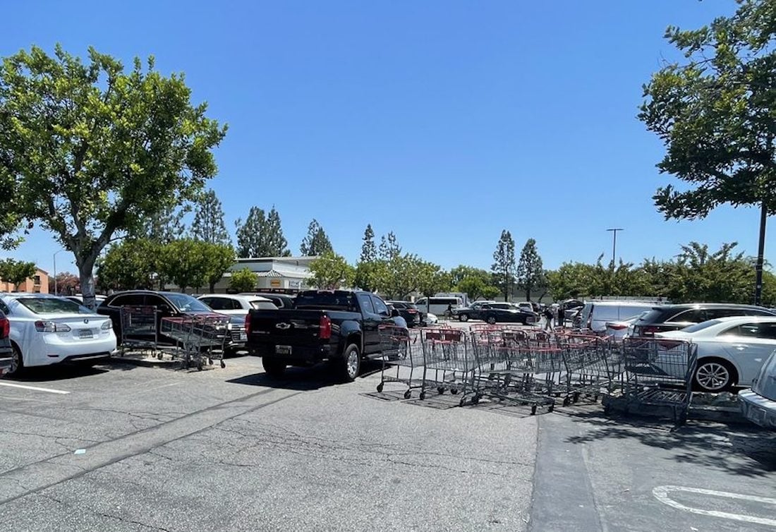 Several shopping carts are scattered and stacked in the middle of a busy parking lot under a clear blue sky, surrounded by parked cars and trees.