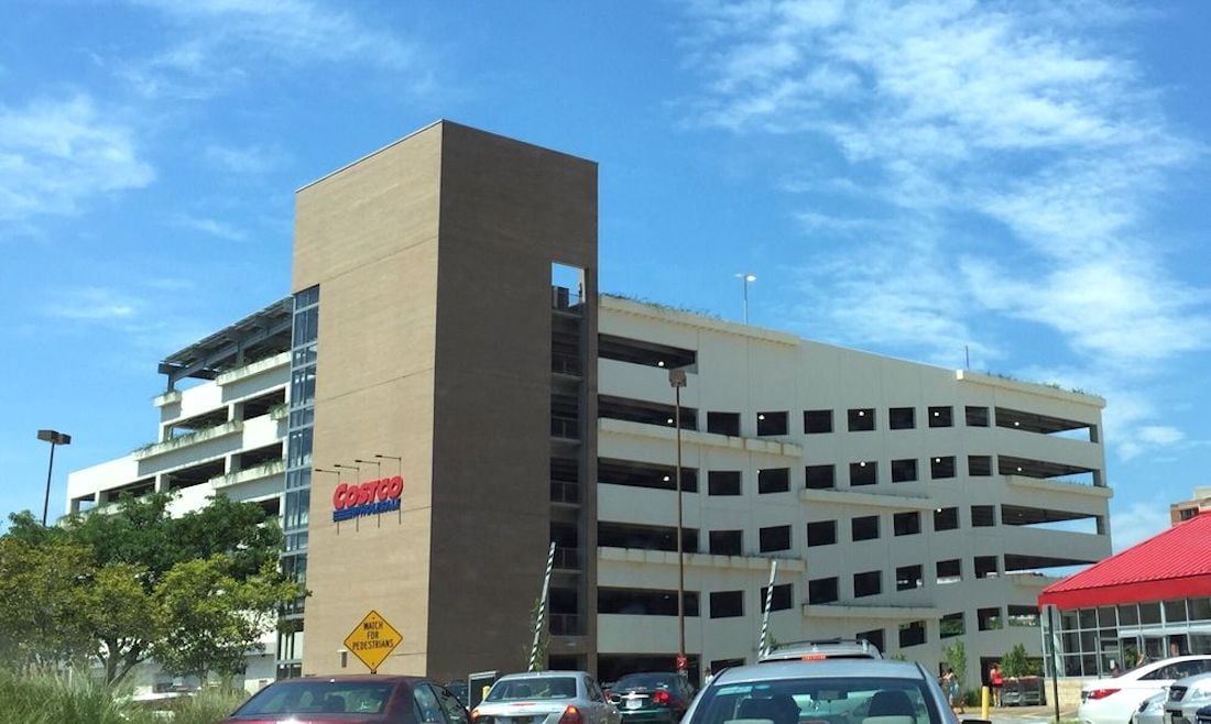 A multi-level Costco parking garage with cars entering and exiting, trees on the left, and a partly cloudy blue sky in the background. A yellow "yield to pedestrians" sign is visible near the entrance.