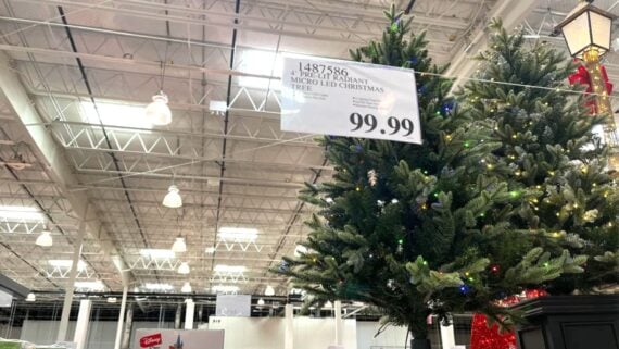 Artificial Christmas tree decorated with colorful lights is displayed indoors under bright warehouse ceiling lights. A sign above the tree reads “4’ PRELIT LED CHRISTMAS TREE” and “99.99.”