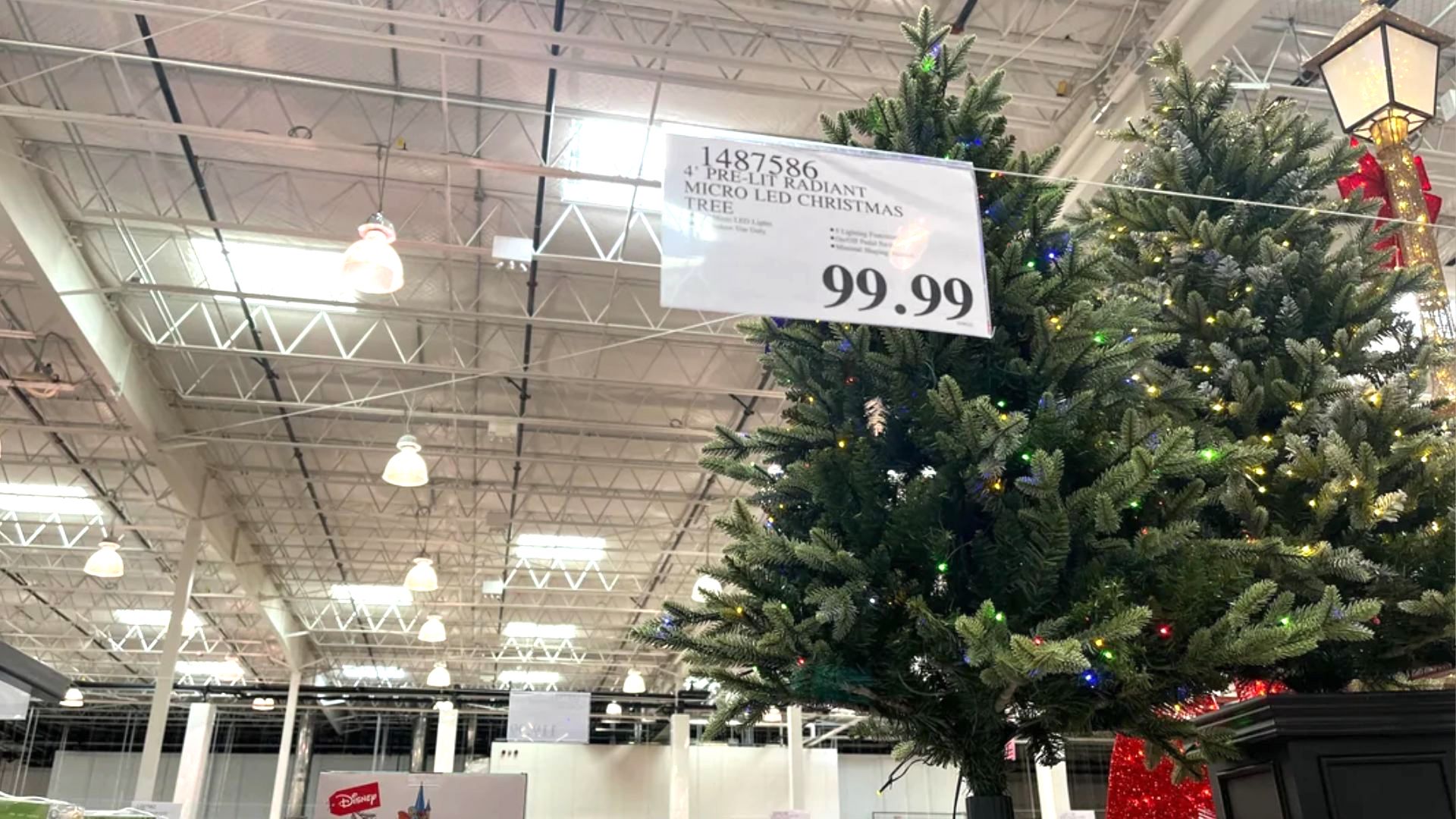 Artificial Christmas tree decorated with colorful lights is displayed indoors under bright warehouse ceiling lights. A sign above the tree reads “4’ PRELIT LED CHRISTMAS TREE” and “99.99.”