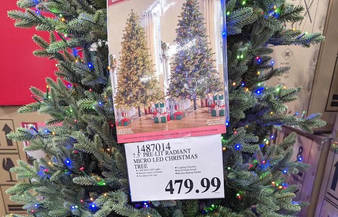 A 7.5-foot pre-lit Christmas tree with multicolored micro LED lights is on display in a store. A sign below lists the price as $479.99, and a product photo shows decorated trees with presents underneath.