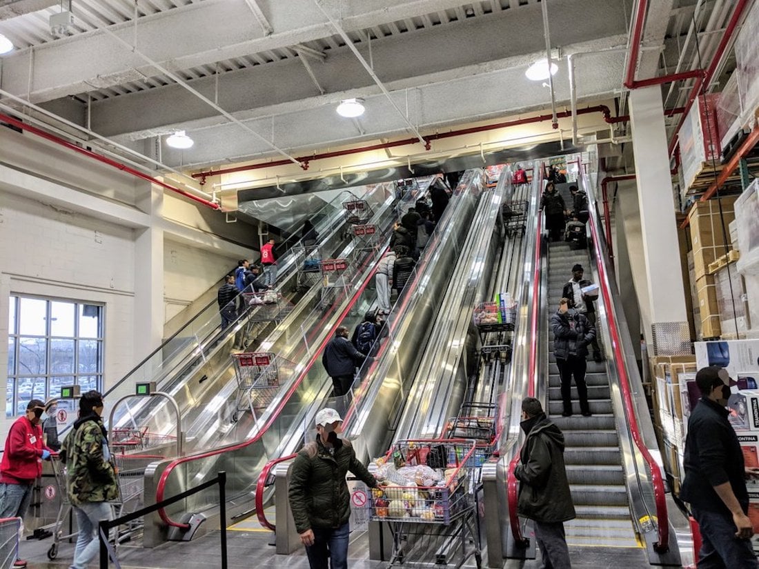 Shoppers with carts ride up parallel escalators in a brightly lit store, while others wait in line at the bottom. Some people wear masks. The scene appears busy and organized.