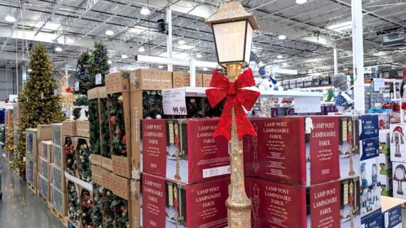 A holiday display in a warehouse store shows stacked boxes of decorative lamp posts, a lit lamppost with a red bow, and Christmas trees with lights in the background. Price tags and other decorations are visible.