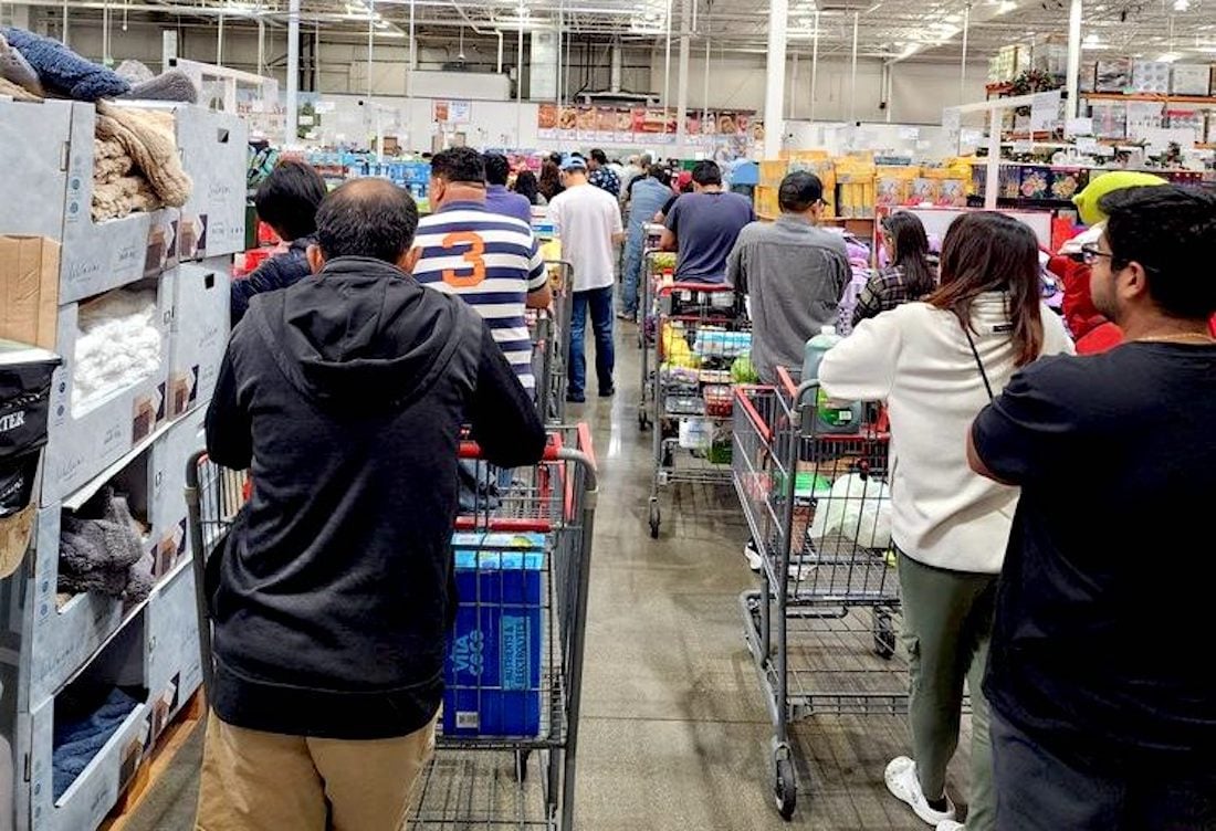 Shoppers with full carts stand in a long checkout line inside a busy warehouse store, surrounded by shelves stacked with various products.