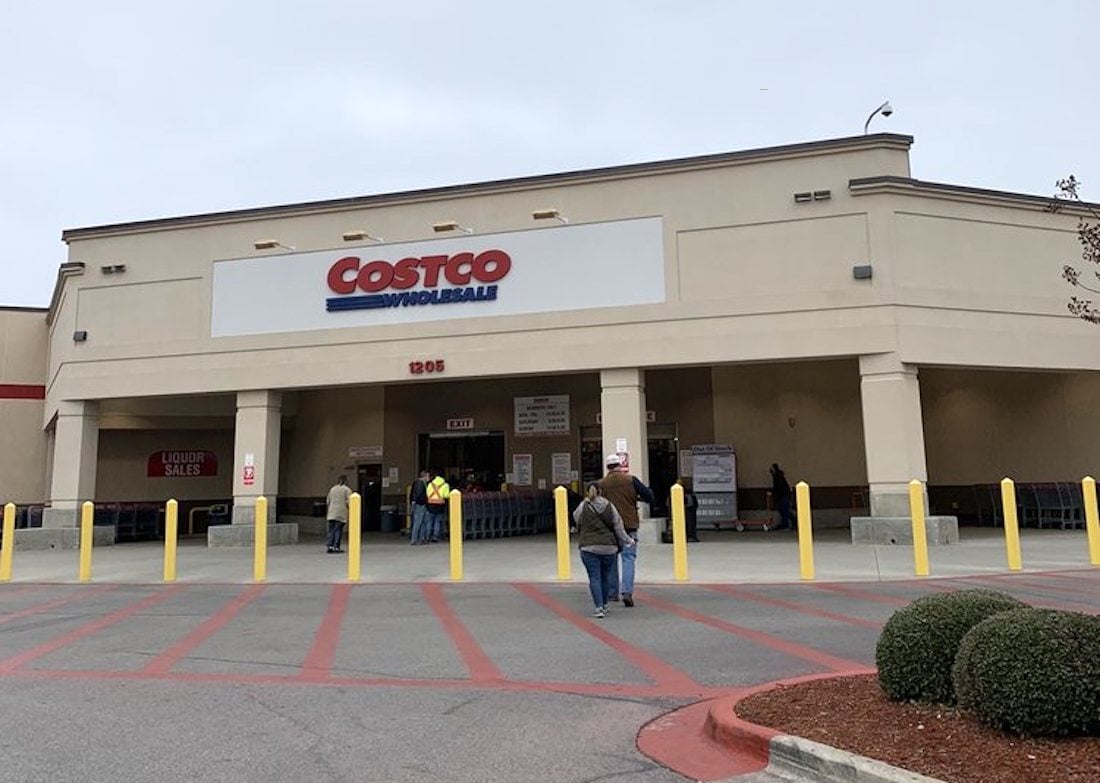 The image shows the exterior of a Costco Wholesale store with people walking toward the entrance. Yellow bollards line the front, and red curbs are visible in the parking area. The sky is overcast.