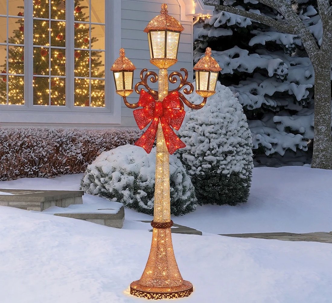 A decorative outdoor lamp post with three lanterns and a large red bow stands in a snowy yard near a house with a lit Christmas tree visible through the window. Snow covers the bushes, ground, and trees.