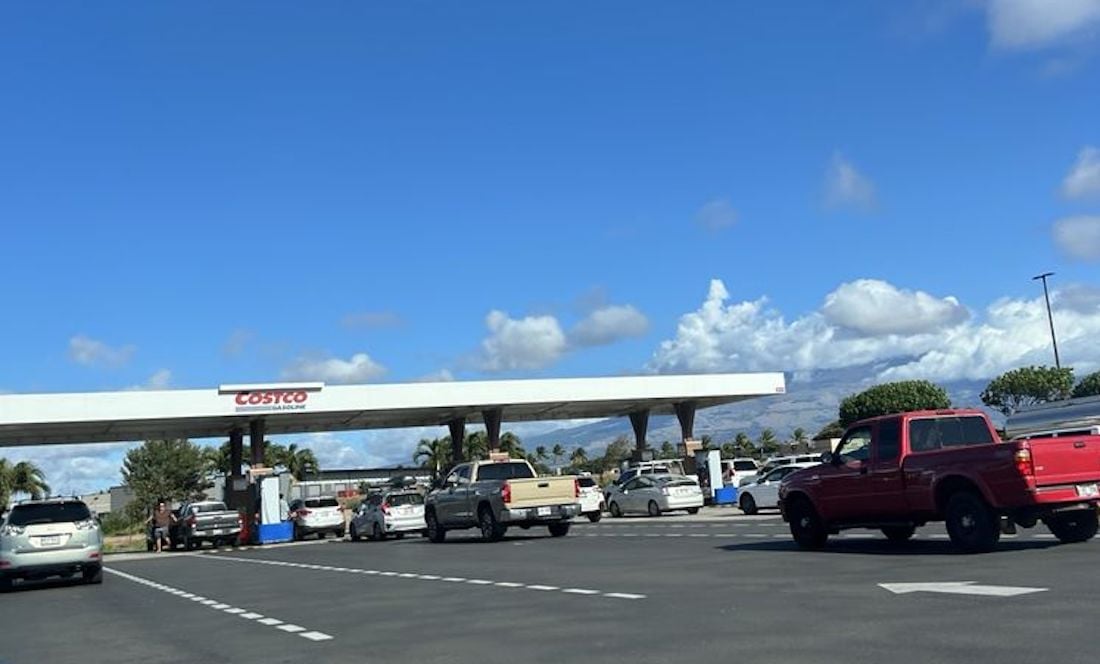 Cars wait in line at a Costco gas station under a clear, blue sky with scattered clouds. The scene includes several vehicles and palm trees in the background.
