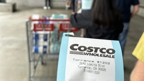 A close-up of a Costco receipt held in front of a shopping cart loaded with bulk items, including water packs, with people standing in line at a warehouse store.