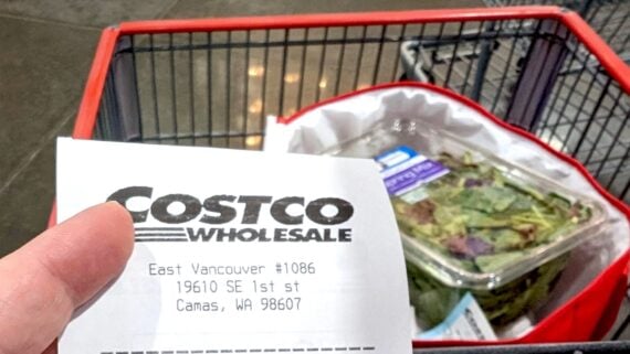 A hand holds a Costco Wholesale receipt in front of a shopping cart with a container of salad greens inside, seen at a Costco store.