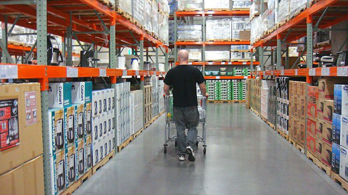 A person pushes a shopping cart down an aisle in a large warehouse store, surrounded by tall shelves stocked with boxes and household items.