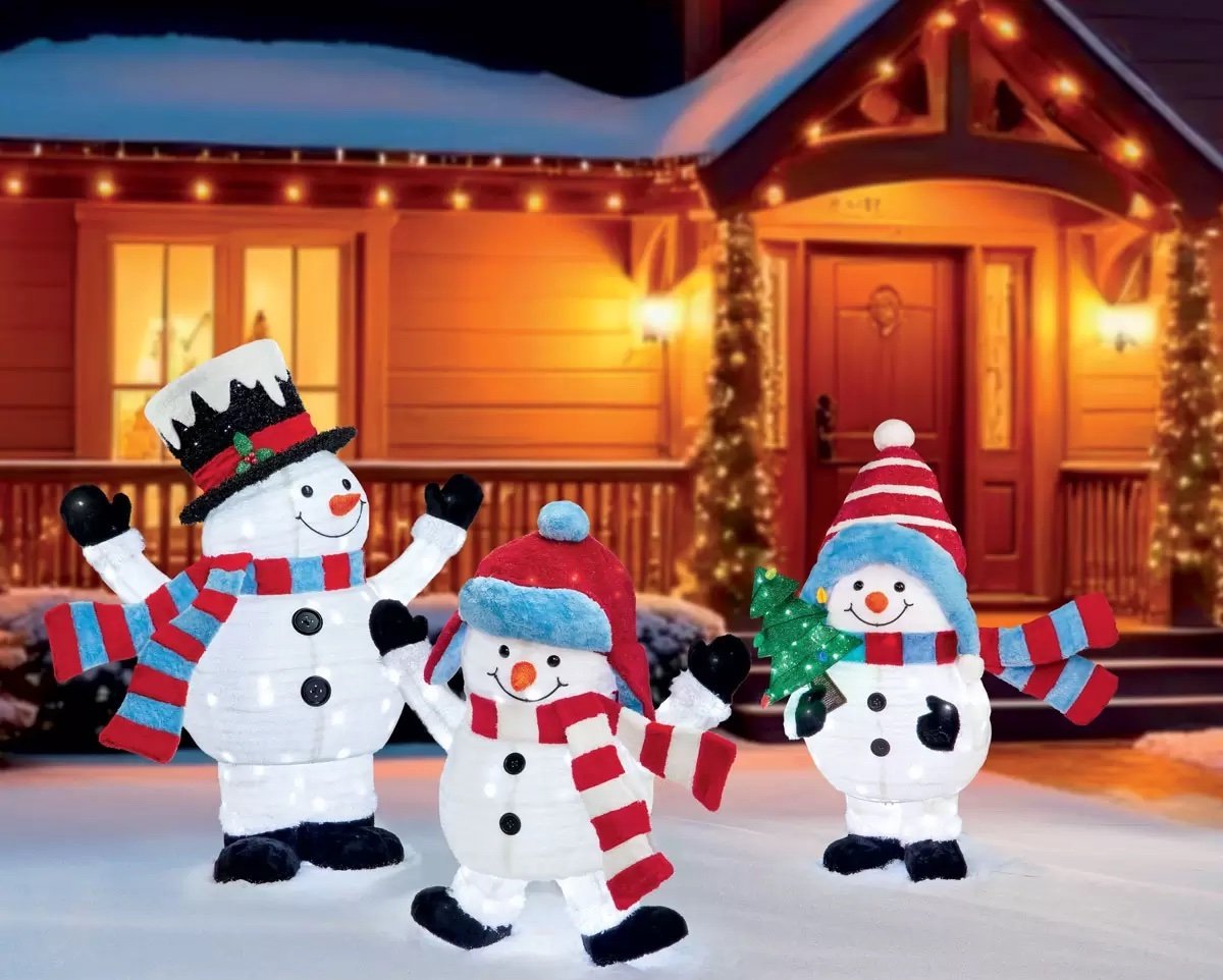 Three light-up snowman decorations wearing hats and scarves stand smiling in front of a house decorated with Christmas lights and a snow-covered yard at night.