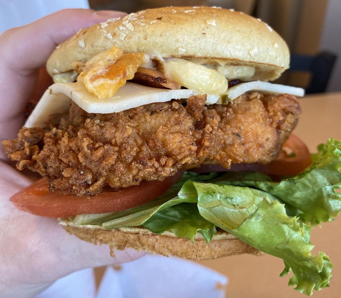 A hand holds a sandwich with a crispy fried chicken fillet, leafy lettuce, tomato slices, white cheese, and bacon on a sesame seed bun.