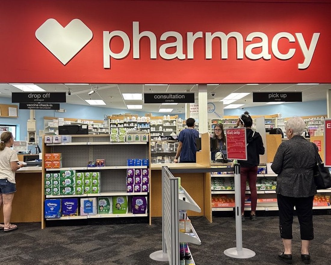 People stand in line at a pharmacy counter inside a store, waiting for service. The red sign above reads "pharmacy" with a white heart logo. Shelves with products and signs for drop-off, consultation, and pick-up are visible.