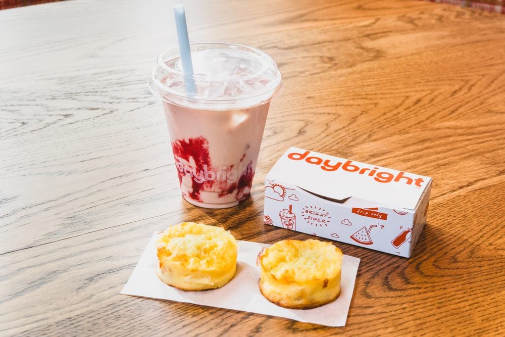 A strawberry milk drink with a straw sits next to a branded “daybright” box and two pieces of buttered English muffin on a wooden table.