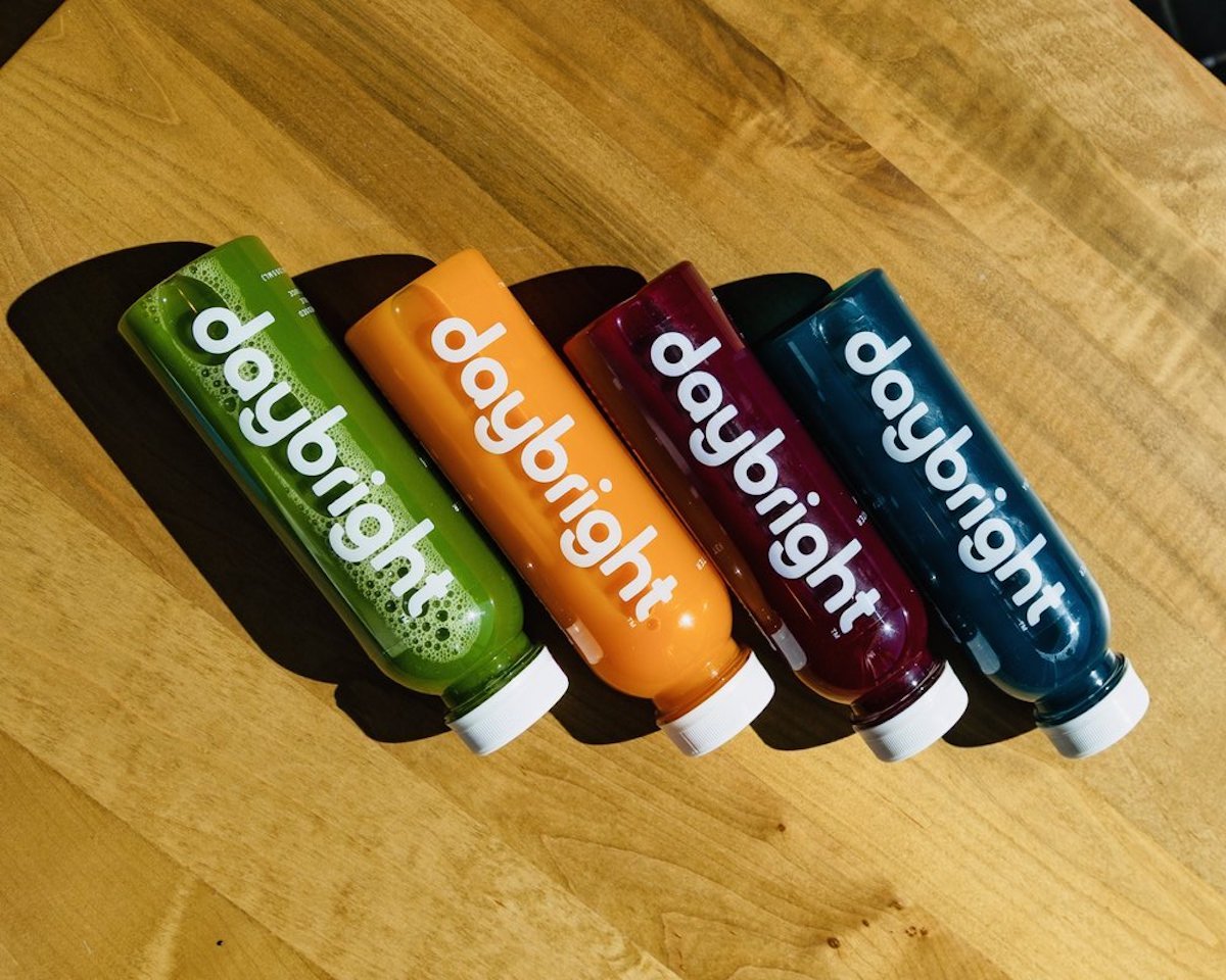 Four bottles labeled “daybright” lie diagonally on a wooden surface. They contain juices in green, orange, red, and dark blue colors, each with a white cap and bold white text on the bottle.