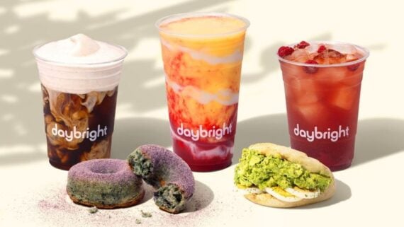Three colorful drinks in clear cups labeled “daybright” are displayed behind two purple matcha donuts and a green tea cream-filled sandwich cookie, all arranged on a neutral surface.